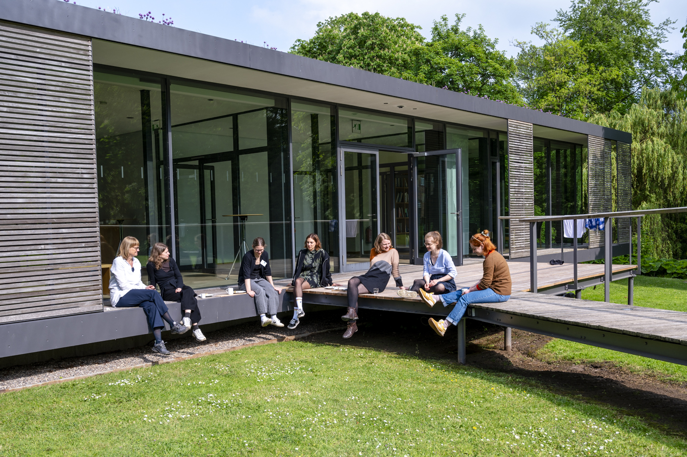 Foto einer Gruppe Menschen, die entspannt auf einer Terrasse in der Sonne sitzen und sich unterhalten.