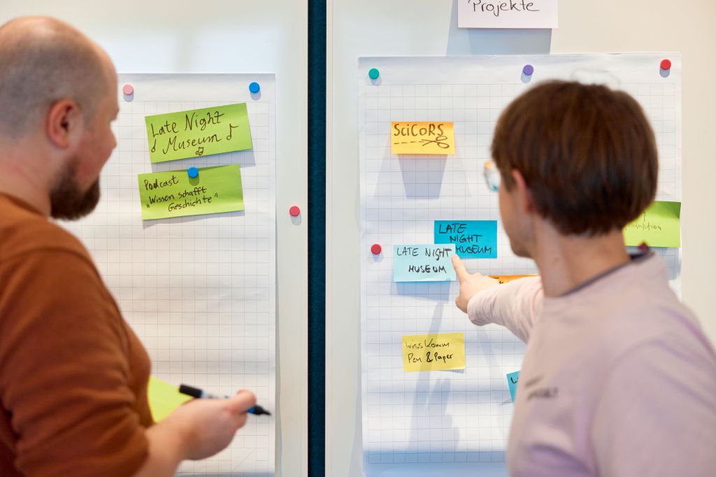 Foto von zwei Männern, die auf ein Whiteboard zeigen, auf dem Post.it kleben.